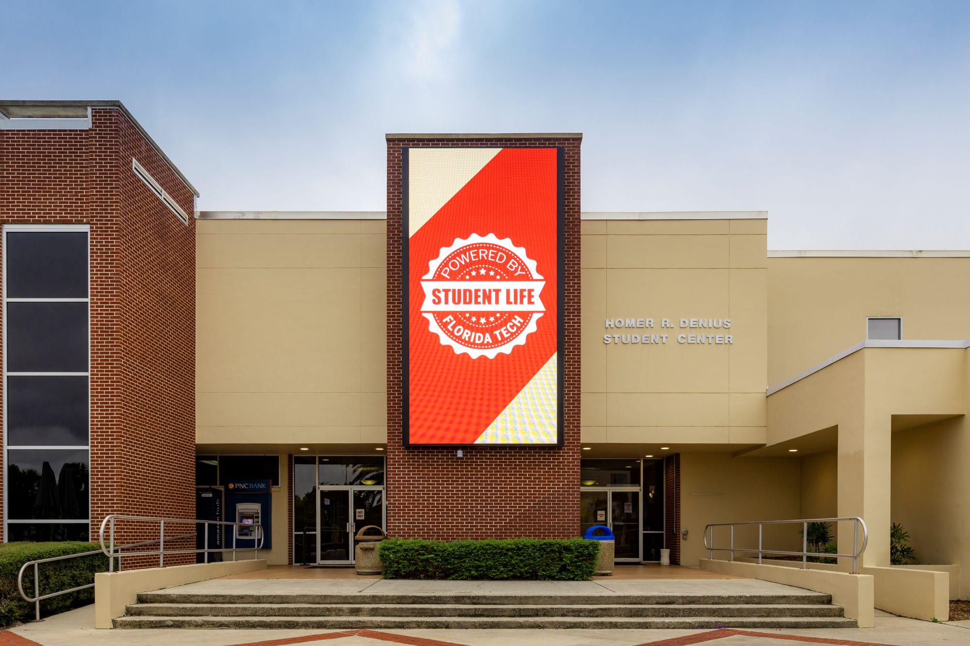 A daytime view of the Homer R. Denius Student Center at Florida Tech. The building features a large digital screen displaying 'Powered by Student Life Florida Tech' in bold red and white colors